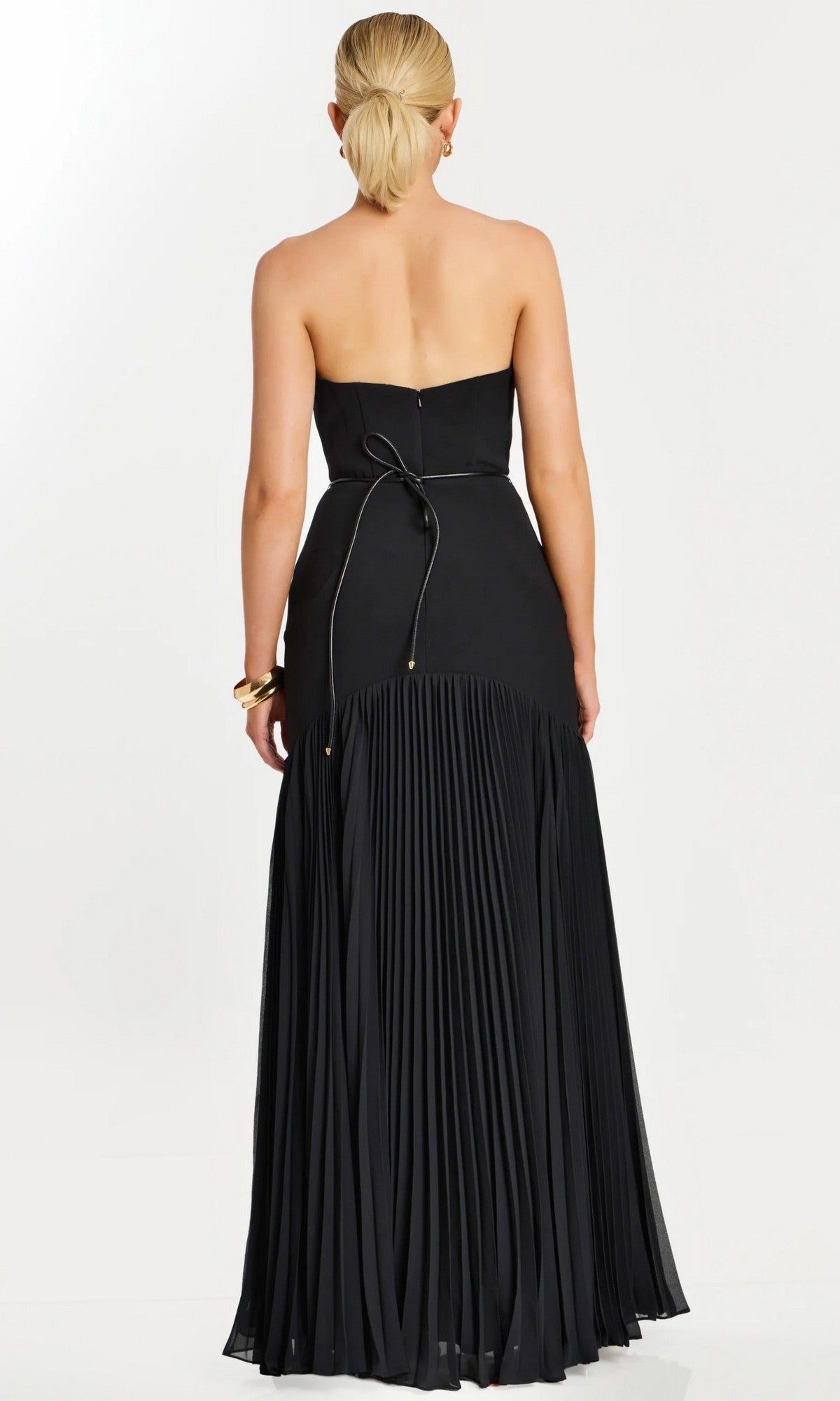 A woman with blonde hair in a low ponytail is pictured from behind against a white background, wearing the Jovani 47000 long formal dress—a strapless black gown featuring a pleated skirt and subtle gold floral detailing at the waist.